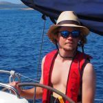 A man at the helm of a sailing yacht wearing a red life vest